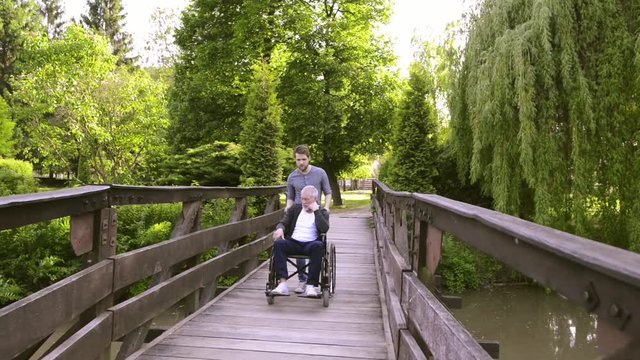 Hipster son walking with disabled father in wheelchair in the park.