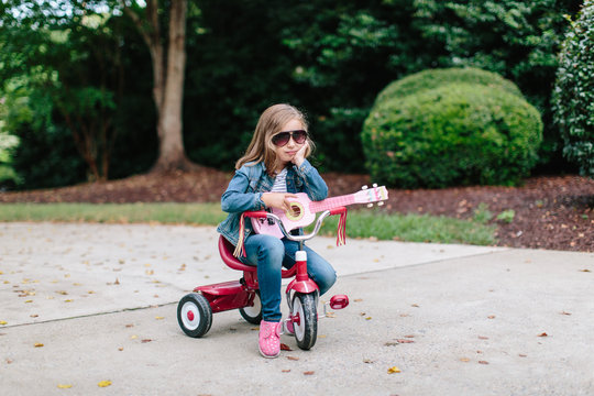 Cute young girl sitting on a tricycle while rocking out with her toy guitar