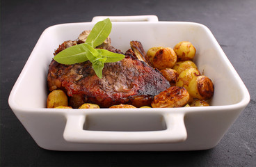 Steak on a bone baked in white dish with vegetables: potatoes, garlic, onions. Black background