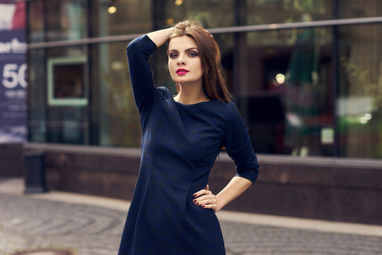 Waist-up portrait of beautiful woman with long brunette hair, wearing dark checkered skater dress with shortened sleeves, standing on city street and posing against large shop windows on background.