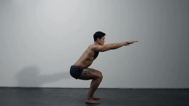 A chiseled fitness model performs squats in front of a grey wall. Long shot. Profile.