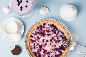Homemade blueberry pie with cream filling served with blueberry smoothie, marshmellow, cookie and milk on pale blue table