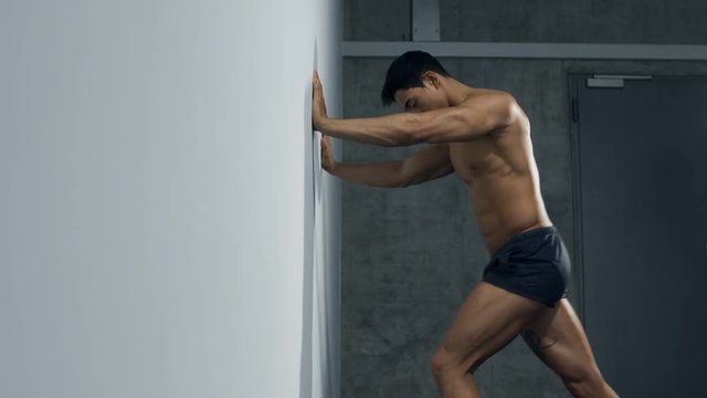 A fitness model stretches his legs pushing himself up against a wall. Profile. Long shot.