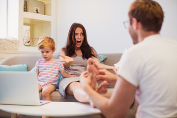 The young man doing foot massage his angry wife who raging him and sitting on the sofa near little son who watching laptop