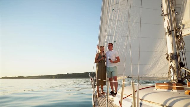 Young man and his wife are sailing river and drinking red wine on a sailboat during the honeymoon. Husband proposing a toast.