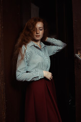 Sensual young red haired model in stripred shirt and red skirt standing near old wall