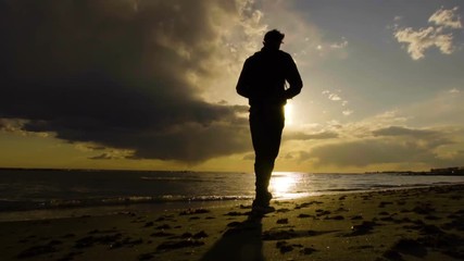 Young man walking on the beach in beautiful sunset at the sea. Cloudy sky, sun setting down to horizon. - Powered by Adobe