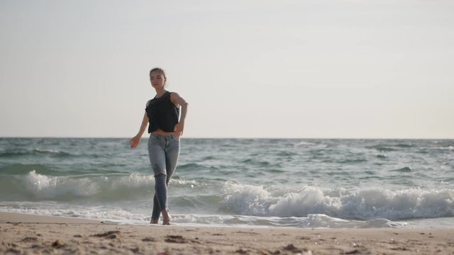 Young woman in casual style - denim and black top doing ballet at the beach. Attractive ballerina practices in hands exercises on sandy plage in autumn. Slow motion. Sea waves background