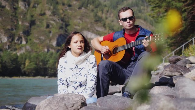 Couple on the beach playing guitar sings song on a summer day next to mountain river. 1920x1080