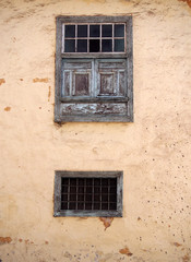 two old wooden windows with ancient faded peeling green paint metal bars shutters and panels on an old plastered peeling concrete terracotta and biege wall with stains and cracks