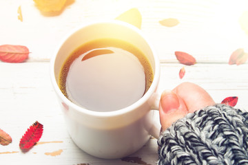 Mug of hot coffee in a woman's hand in a sweater in the autumn setting on a wooden table with a knitted scarf, sweater. Comfort, warmth, cozy.