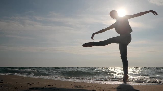 Young woman in casual style - jeans and black top dancing ballet at the beach. Attractive ballerina practices in stretching on sandy coastline in autumn. Slow motion. Beautiful scene. Gimbal shot