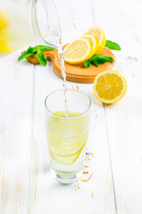 In a glass beaker, a cold lemonade is poured from a jug on a white wooden background surrounded by lemons.