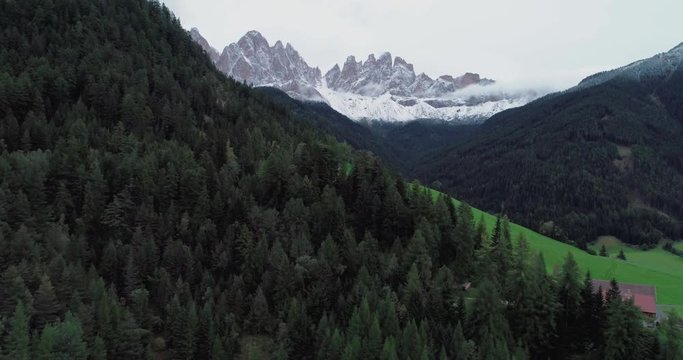 Amazing aerial shot of Dolomiti mountain on a cloudy autumn day. Flying above beautiful green forest in Santa Maddalena in Val di Funes, Dolomites, Italy with view over Italian Alps.