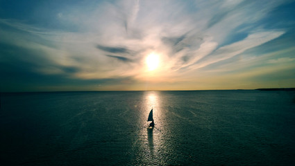 Little white boat floating on the water towards the horizon in the rays of the setting sun. Beautiful clouds with yellow highlights. Aerial view - obrazy, fototapety, plakaty