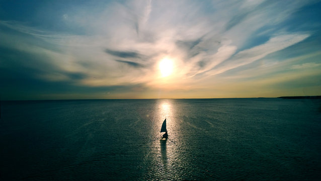 Little white boat floating on the water towards the horizon in the rays of the setting sun. Beautiful clouds with yellow highlights. Aerial view