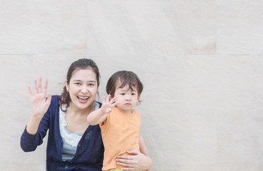 Closeup asian mother and son waved hand to greeting someone in happy motion on marble stone wall textured background with copy space