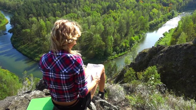 Young blonde woman tourist on a cliff over river Berd studying a map and view the neighborhood. Russia, Siberia, Salair
