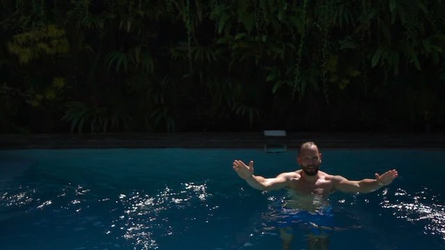 Man standing in the swimming pool and splashing water, slow motion shot at 240fps, steadycam shot 

