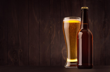 Brown beer bottle and glass weizen with golden lager on dark wood board, copy space, mock up. Template for advertising, design, branding identity.