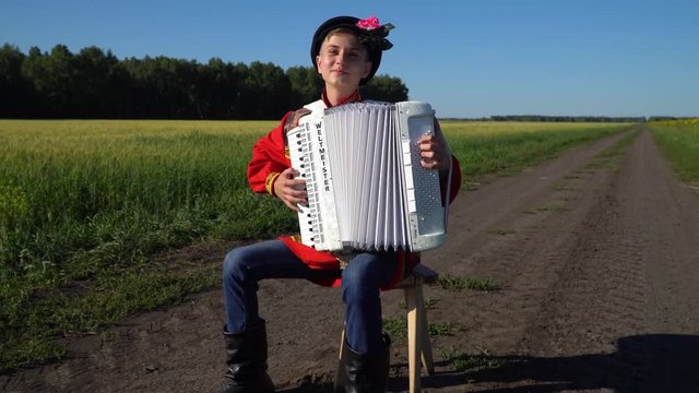 A young man in a Russian national traditional costume plays the accordion sitting on a chair on a country road, among a wheat and sunflowers field. Forest on the background.