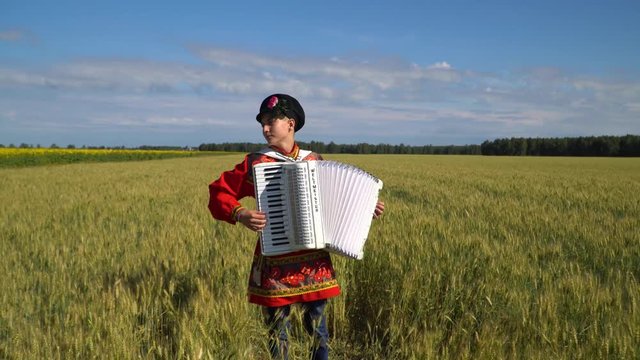 A young man in a Russian national traditional costume standing in a wheat field and playing the accordion.
