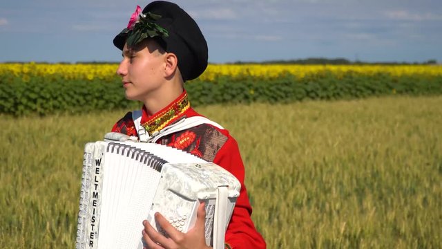 A young man in a Russian national traditional costume plays the accordion in the wheat field.