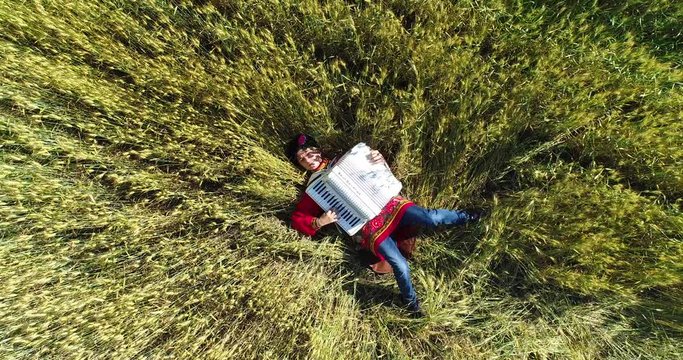 Aerial: A young man in a Russian national traditional costume plays the accordion lying in a wheat field.
