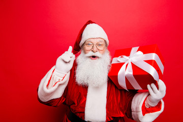 Ho ho! Holly jolly x mas! Noel magic! Funny kind astonished saint nicholas in traditional head wear is presenting a wrapped gift with white tape bow with forefinger up, isolated on red background