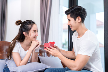 Young asian couple together, man holding surprise box heart gift to woman on sofa in home at living room relax and resting happy lover romantic sweet interior indoor concept.