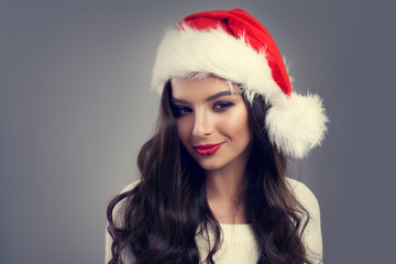 Christmas Portrait of Beautiful Smiling Woman in Red Santa Hat. New year Fashion Model Brunette with Makeup and Healthy Wavy Hair