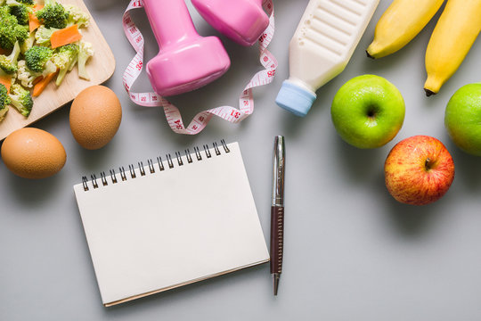 Healthy eating, dieting, slimming and weight loss concept - Top view of green apple, bananas, measuring tape, dumbbells, blank note book and vegetables on grey background. copy space for Text