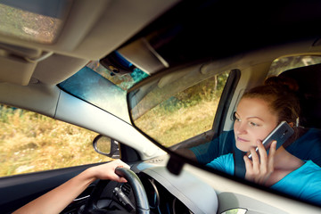 Reflection in the mirror of beautiful young woman driving a car and talking on smart phone, free space. Business woman sitting in car with cell phone, looking on road