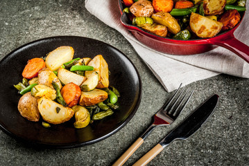 Plate and Skillet with  fried seasonal autumn vegetables (zucchini, potatoes, carrots, beans), on black stone table copy space