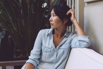 Beautiful model look asian woman with dark hair and smooth tanned skin wearing blue jean dress is looking aside while sitting on a white sofa in a modern city coffee shop on a plants background.