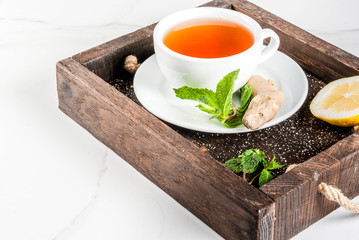 Cup of ginger tea with lemon,fresh mint and honey, in wooden tray, on white marble table, copy space