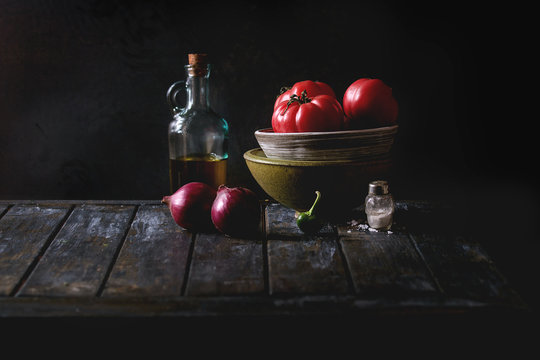 Bowl of tomatoes with purple onion, pepper and bottle of olive oil on old wooden kitchen table. Dark rustic still life.
