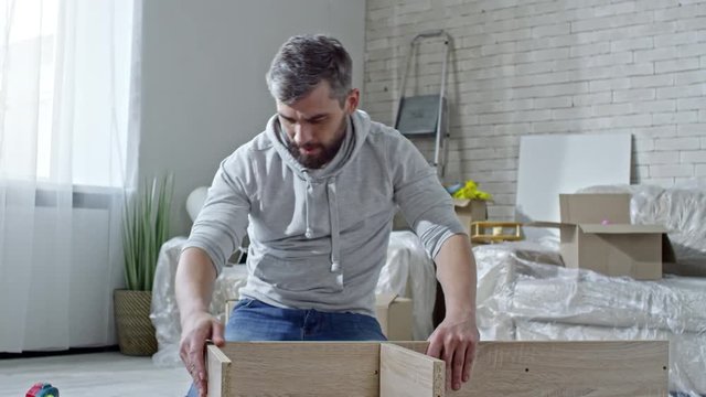 PAN of man with beard and grey hair sitting on floor of new apartment and assembling wooden shelf; cardboard boxes and sofa covered in plastic wrap standing in background