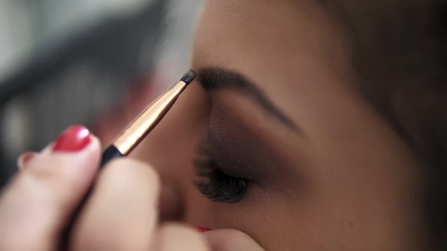 Closeup view of the makeup artist's hands using brush to paint eyebrows for a model with false lashes. Slowmotion shot
