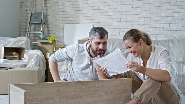 PAN of laughing man with beard and his beautiful wife sitting on floor of new apartment and looking at assembling manual for wooden shelf