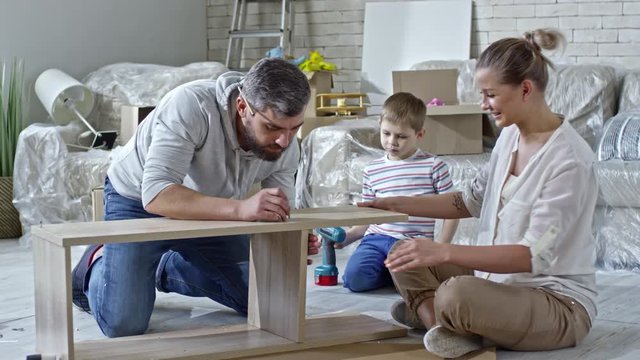 PAN of concentrated man with beard putting screws into wooden pieces of shelf as his beautiful wife helping him; little boy with toy drill sitting beside