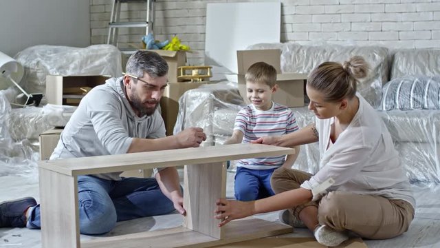 PAN of beautiful woman sitting on floor of new apartment and holding wooden pieces of shelf while her husband putting screws into it; cute little boy helping dad