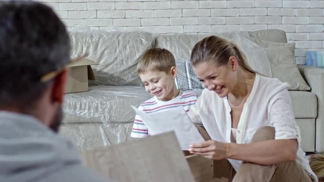 Laughing young woman sitting on floor of new apartment and looking at furniture assembling manual, then discussing it with husband showing her wooden pieces; cute little boy sitting beside