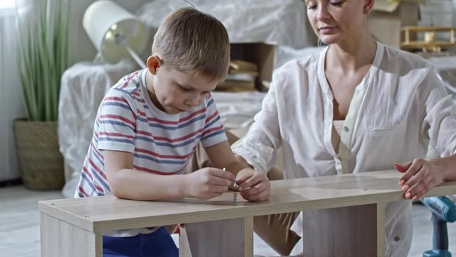 Tilt up of little boy driving screws into wooden shelf while young beautiful mother helping and smiling