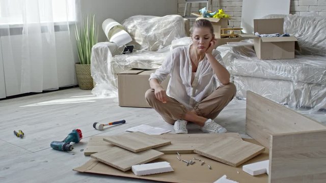 PAN of confused young woman sitting of floor of new apartment and reading assembling manual, then looking at wooden pieces of furniture lying before her and thinking