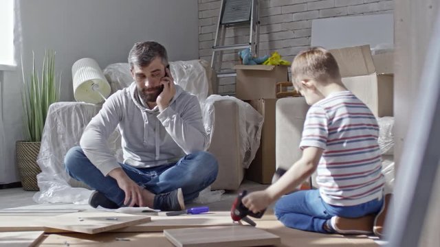 PAN of little boy sitting on floor of new house and playing with toy construction tools while his bearded father talking on mobile phone and smiling