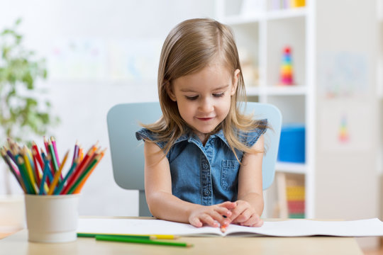 Little kid girl draws sitting at table in room in nursery