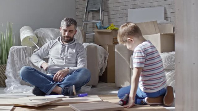 PAN of bearded man sitting on floor of new house and talking on mobile phone as little boy playing with disassembled furniture and toy tools; armchairs in plastic wrap and boxes in background
