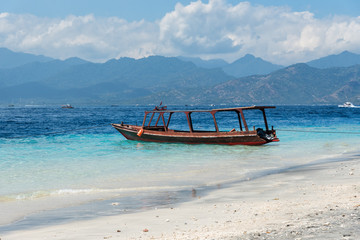 Small wooden boat on blue beach with cloudy sky and Lombok island on background. Gili Trawangan, Indonesia.