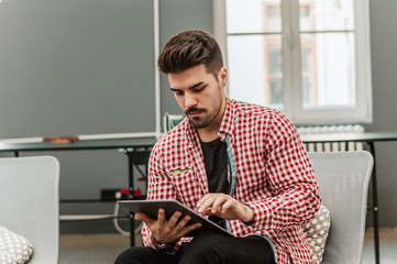 Young male in casual wear browsing internet on digital tablet while sitting in his home.